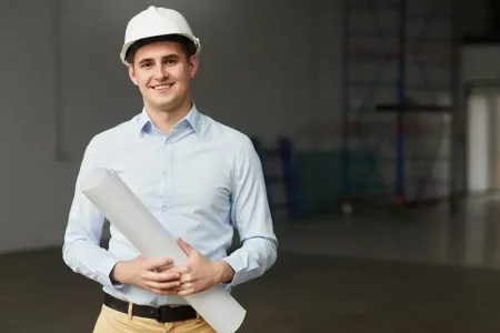 portrait-young-architect-work-helmet-holding-blueprint-smiling-while-standing-empty-building